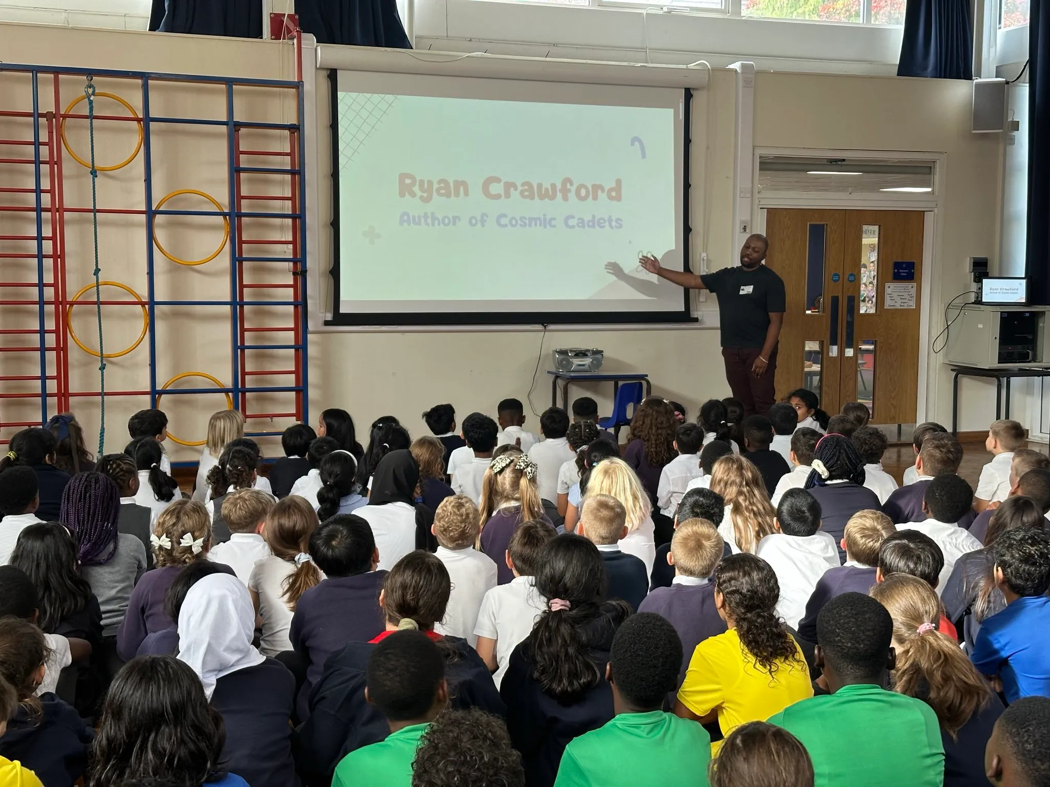 Ryan Crawford presenting to a full school hall of seated pupils, his Cosmic Cadets title slide on the screen behind him