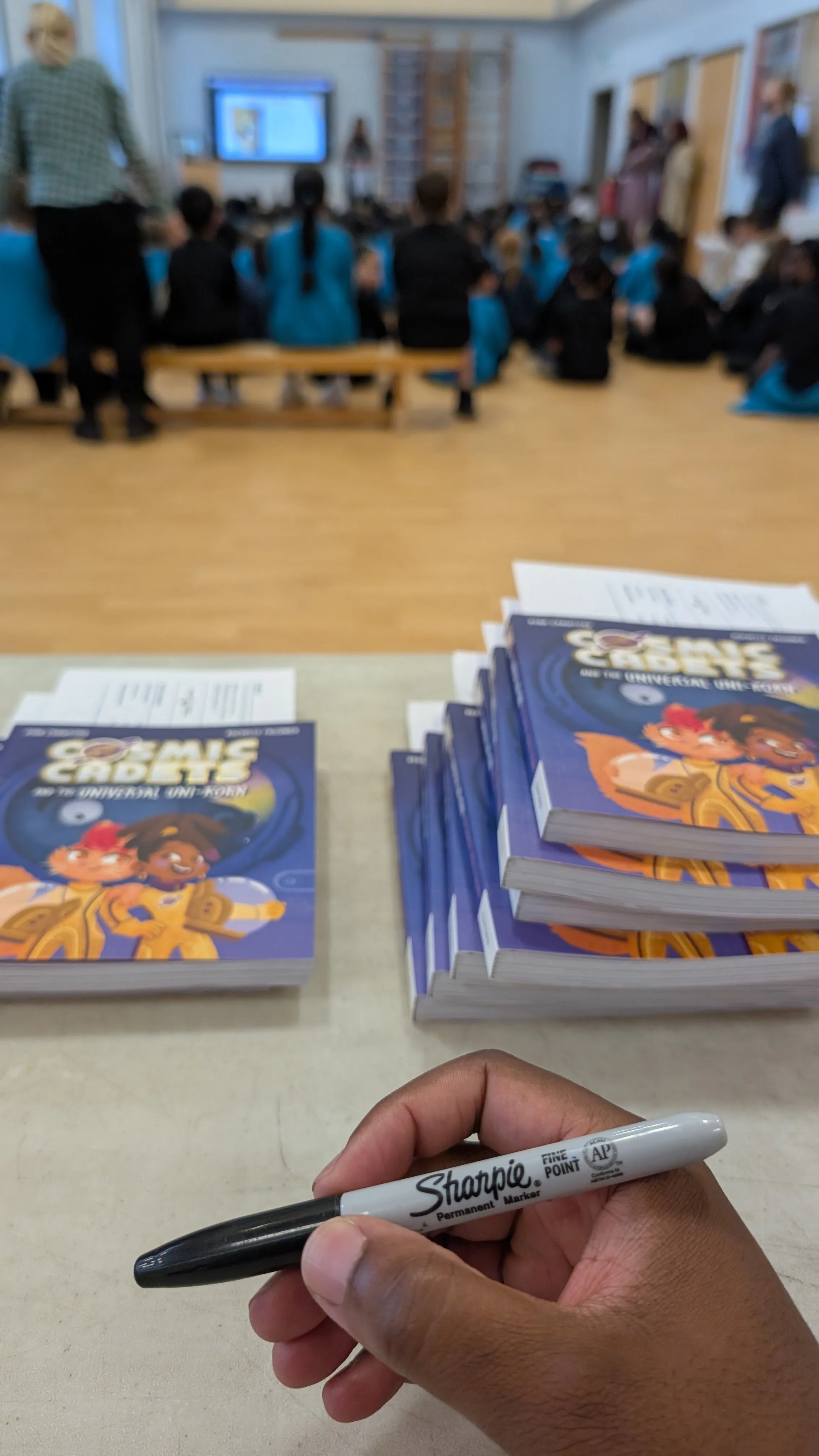 A Sharpie held over stacks of Cosmic Cadets paperbacks, with a school hall of pupils out of focus behind